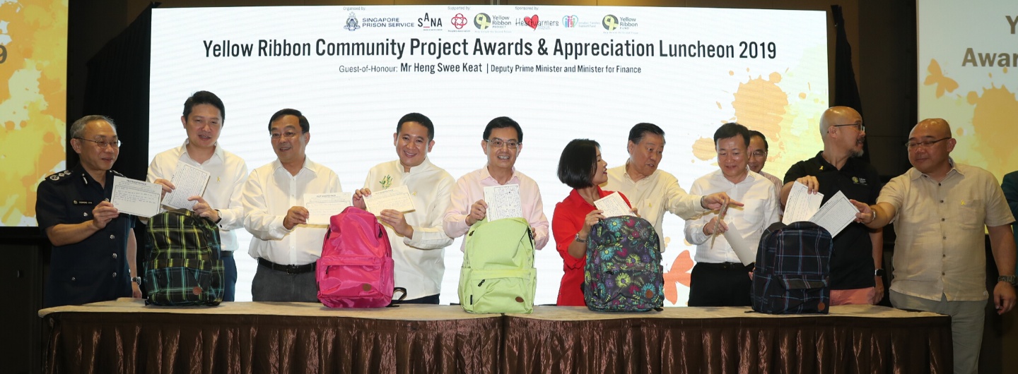 People at Yellow Ribbon event holding papers behind backpacks on a draped table.